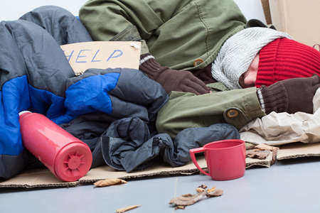 Homeless man sleeping on cardboard with sleeping bag and thermosの写真素材