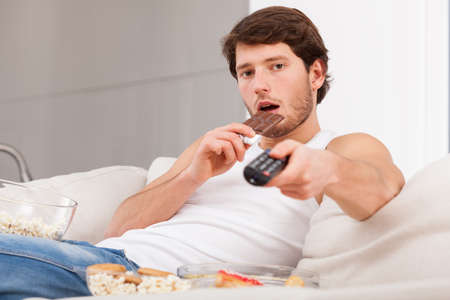A man eating a chocolate bar holding a remote control on a couchの写真素材