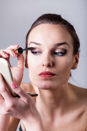 Woman painting eyelashes using mascara on isolated grey background