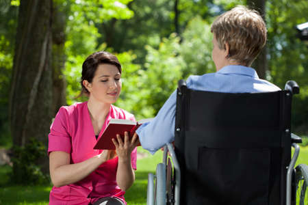 Caregiver reading a book to disabled senior woman outdoors