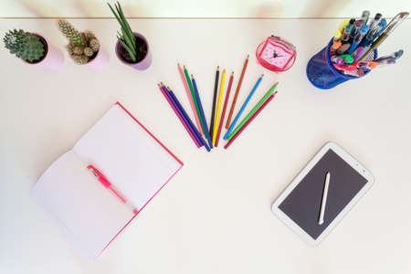 Top view of open exercise book, pencil crayons and tablet on white desk