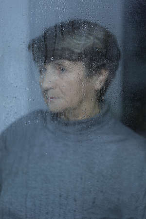 Photo of older sad woman behind the rainy glass