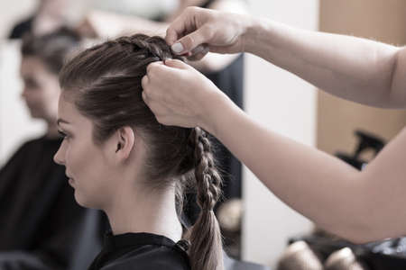 Female hairdresser is braiding young woman's hair