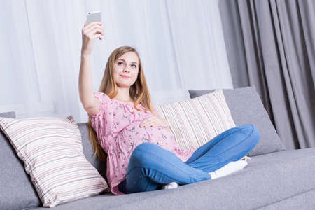 Smiling girl in pregnancy, sitting on sofa, taking selfie with cellphone