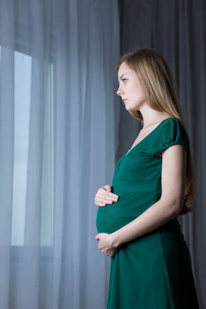 Worried pregnant girl standing in front of window, touching her tummyの写真素材