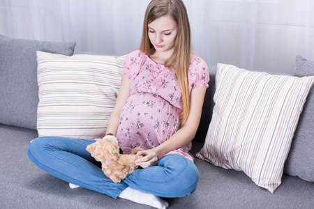 Girl in pregnancy holding teddy bear, sitting on sofa