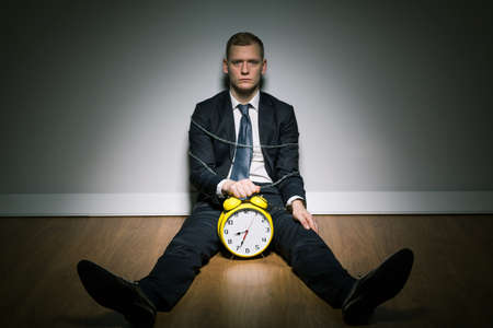 Man sitting on floor in dark interior, with alarm clock between his legs and chain round his body.の写真素材