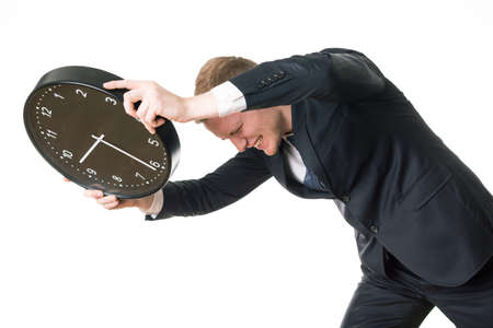 Angry man in suit holding black clock, trying to break it, standing on white background.