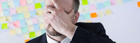 Overworked corporate worker covering face with hand, standing on sticky notes wall background, panoramaの写真素材