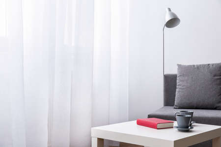 Book and two cups lying on small table. Room with white curtain, lamp and grey sofa.の写真素材