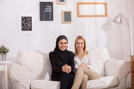 Happy muslim and european, blonde woman sitting together on sofa in light, cozy interiorの写真素材