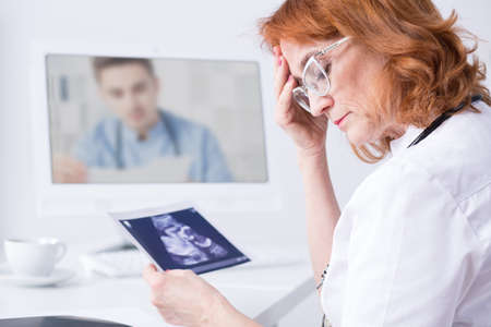 Pensive, mature woman doctor holding ultrasound, picture during video consultation with another medic, sitting in light officeの写真素材