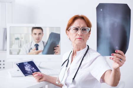 Mature woman doctor analysing ultrasound picture during medical consultation with another doctor via...