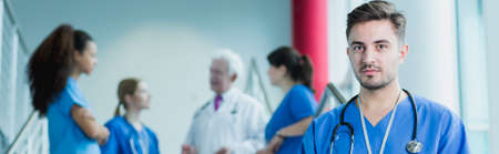 Young handsome future physician on his internship in hospital. Behind him colleagues with older...