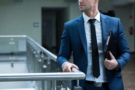 Elegant man leaning against railing in modern, office building, holding folder with documents