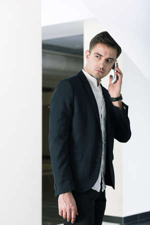 Young student in suit talking on cellphone, standing in light interiorの写真素材