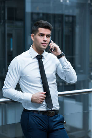 Elegant man leaning against railing in modern, office building, talking on cellphone