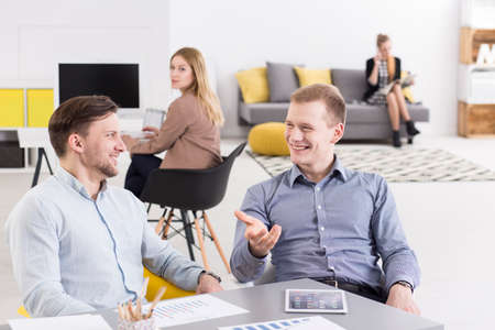 Two men smiling, working together in light office, in the background two elegant women