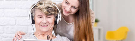 Mature woman with headphones and young, beautiful girl sitting together in light interior, panoramaの写真素材