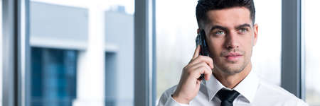 Young handsome busy businessman controlling his business cases by mobile phone. Standing in officeの写真素材