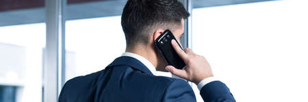Elegant young man in suit talking the phone in his modern office. Standing back to the photoの写真素材