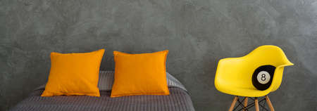 Close-up of a bed covered with grey bedspread, with two orange pillows on top and a yellow designer chair besideの写真素材