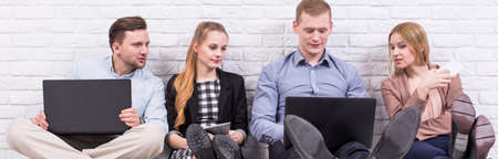 Panorama of four colleagues sitting on the floor by a white brick wall, working at their computersの写真素材