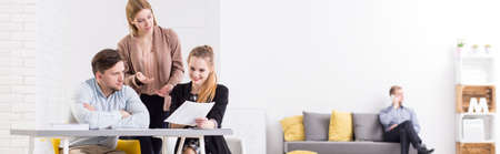 Panorama of a modern, minimalist office with team of colleagues discussing documents at a white desk and a young man talking on the phone in the backgroundの写真素材