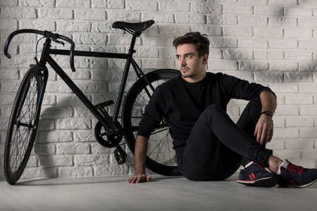 Man sitting on floor next to a black bike, brick wall in the backgroundの写真素材