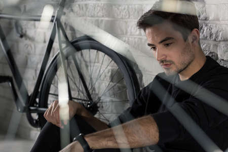 Sad man sitting on floor next to bicycle, brick wall in the backgroundの写真素材