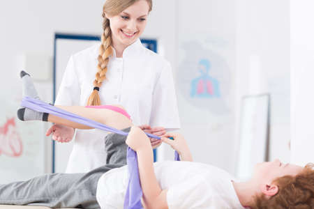 Little patient exercising his leg using elastic bend during physical therapy. Next to him smiling young physiotherapistの写真素材