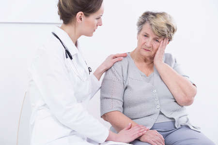 Worried senior patient and positive doctor with stethoscope sitting in light interiorの写真素材