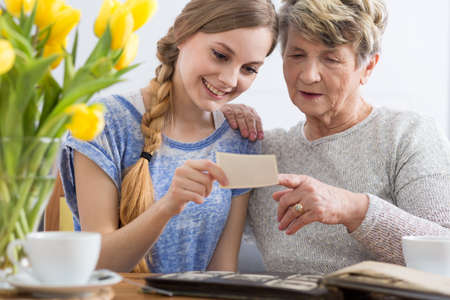 Young girl and grandmother watching together photo album, sitting in cozy interiorの写真素材