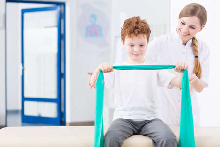Little red head boy exercising with a elastic band in physical therapy clinic. Next to him young...