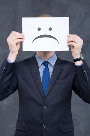 Shot of a man in a suit holding a paper with a sad face on itの写真素材