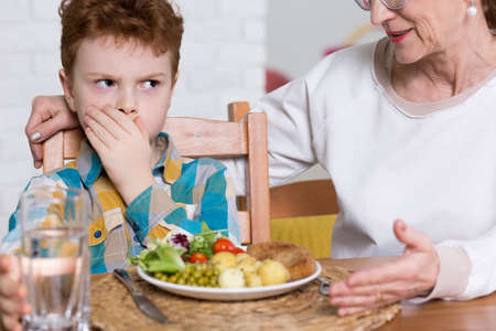 Young naughty boy refusing to eat healthy dinner with vegetables. Next to him his grandmother