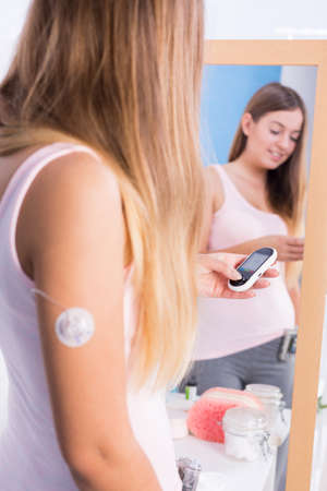 Woman with diabetes standing in front of big mirror, holding glucometer, smiling.の写真素材