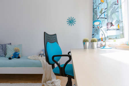 Bright teenager room arranged in white and blue, with large white desk and a swivel chair beside a large windowの写真素材