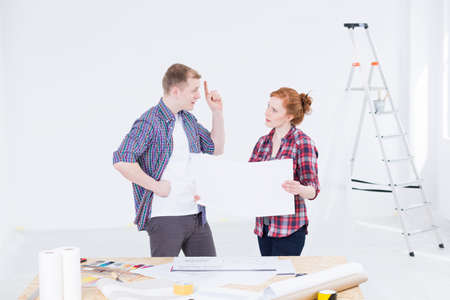 Young man and woman in work clothes discussing a technical drawing inside a room under renovationの写真素材