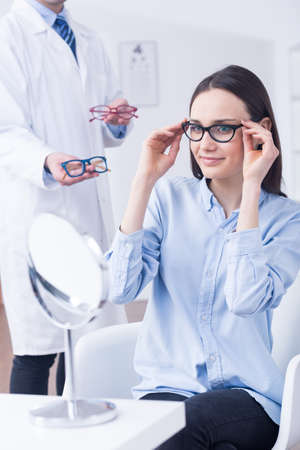 Attractive woman trying on black frames, optician holding red and blue farmes, light interiorの写真素材