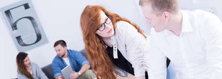 Pretty young red head girl talking with her handsome colleague at work. Behind  them man and woman...