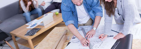 Two young people working together on a new building project in company. Leaning above paper with...