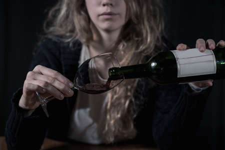 Shot of a young blonde woman sitting at the table and pouring herself a glass of wineの写真素材