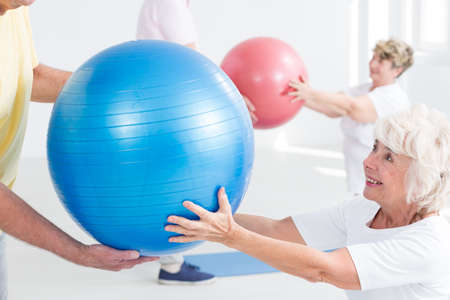 Close-up of a cheerful elderly lady passing a blue exercise ball to her fitness partnerの写真素材