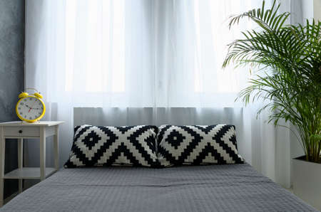 Modern bedroom with grey bed. On the top black and white pillows. Next to bed yellow clock alarm on nightstandの写真素材
