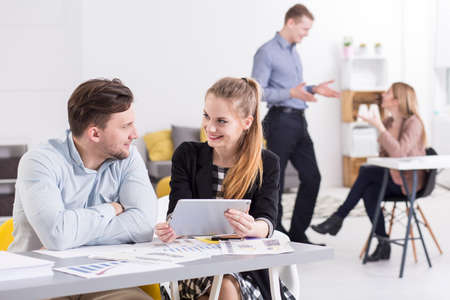 Pretty young smiling woman working with handsome young man in office. In the background two...