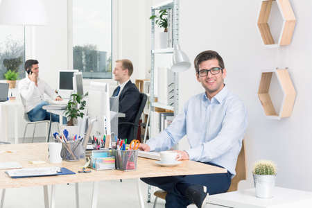 Happy designer sitting beside desk in modern studio, in the background two men discussing