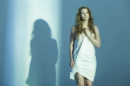 Young, bruised woman dressed in white, standing in an empty room where she casts a shadow on one of the wallsの写真素材