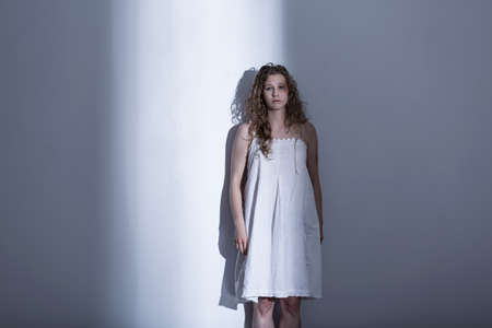 Young, brused woman in a nightdress, standing by a white wall in an empty roomの写真素材