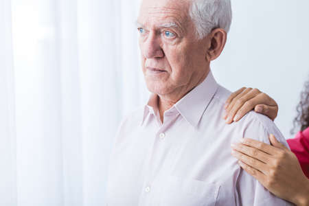 Sad senior man and woman's hands on his arms, light interior.の写真素材